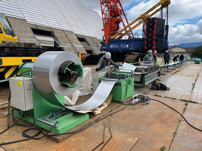 地上で屋根材を現場成型。写真手前からアンコイラーでコイルを回し、カットして、成型作業を行う